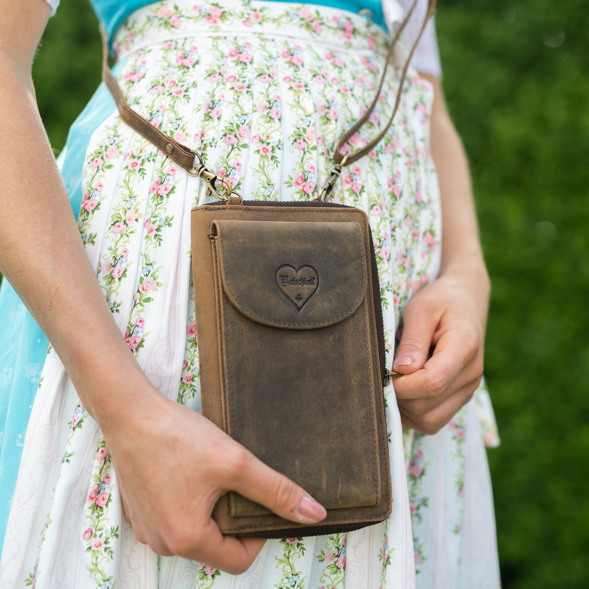 Frau trägt braune Hodalump Leder-Handytasche mit Herzlogo über floralem Dirndl.